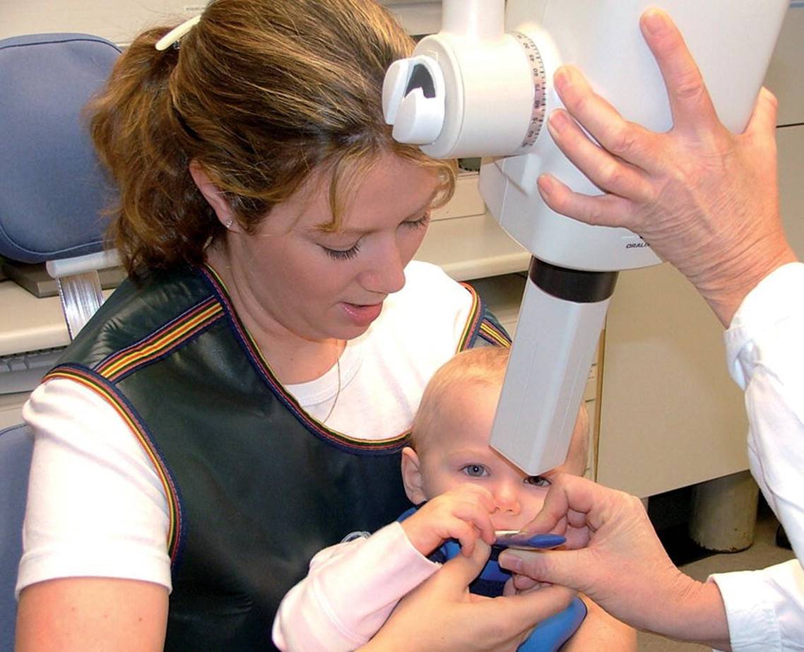 Photo displaying a toddler being assisted by a woman for dental examination.